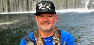 Man by a waterfall wearing a fishing hat and gear, enjoying fishing time related to Buck-eye.