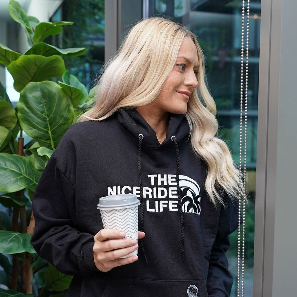 Woman wearing a black hoodie with 'THE NICERIDE LIFE' design, holding a coffee cup, showcasing stylish and comfortable hoodies.