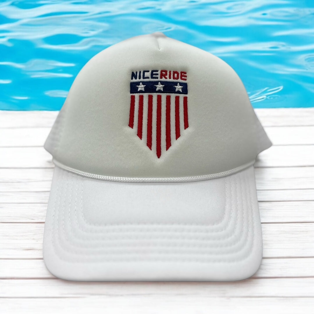 NICERIDE Goal Foam Trucker Hat in front of blue water, featuring embroidered red and blue shield design.