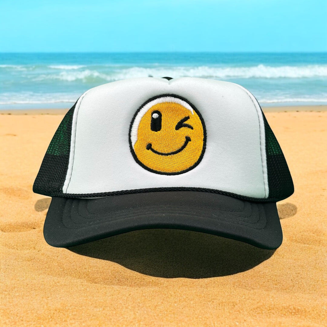 Wink Foam Trucker Hat with smiley face on a beach, perfect for summer style and comfort.