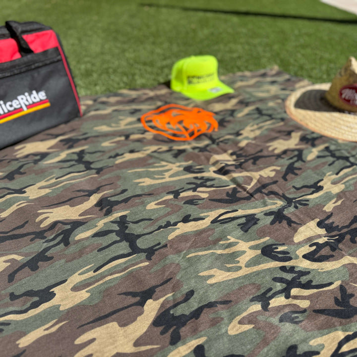 Niceride Palm camouflage fleece blanket spread out with orange logo, hat, and cooler bag on grass surface.