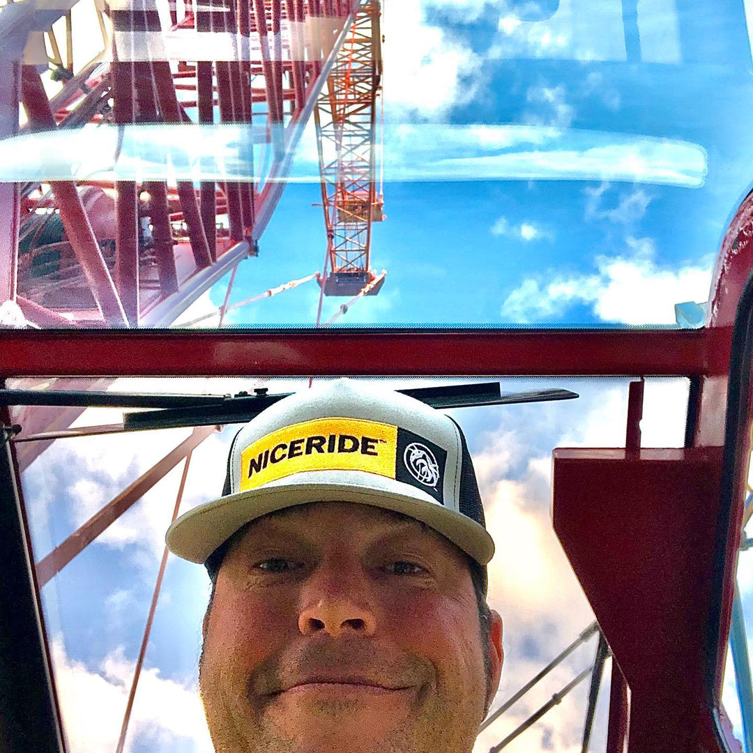 Selfie of man wearing Dozer trucker hat, with construction crane in the background under a blue sky.