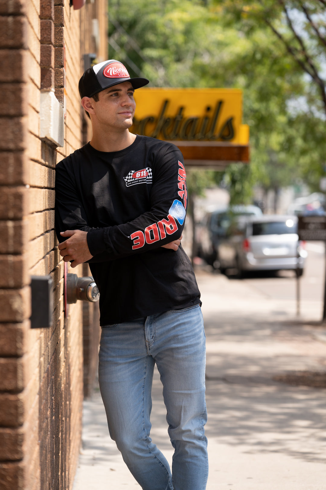 Man wearing a 619 Motor Speedway long sleeve cotton t-shirt with blue jeans, leaning against a wall outside.