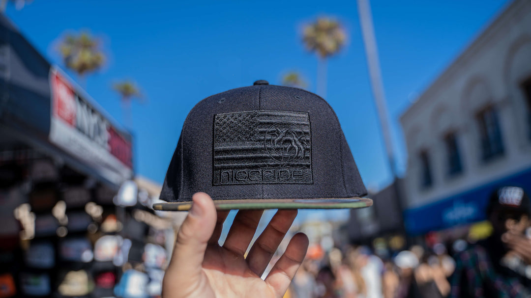 NICERIDE Strong - BLACK Snapback Hat featuring American flag embroidery, held in hand against a sunny marketplace backdrop.