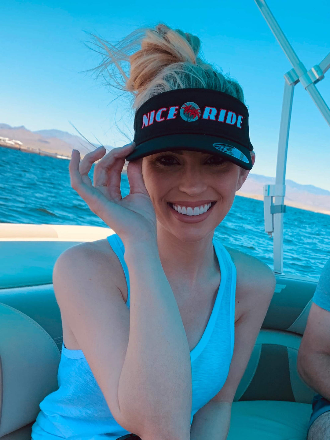 Smiling woman wearing a black visor, showcasing the NICERIDE design, on a boat with a scenic background.