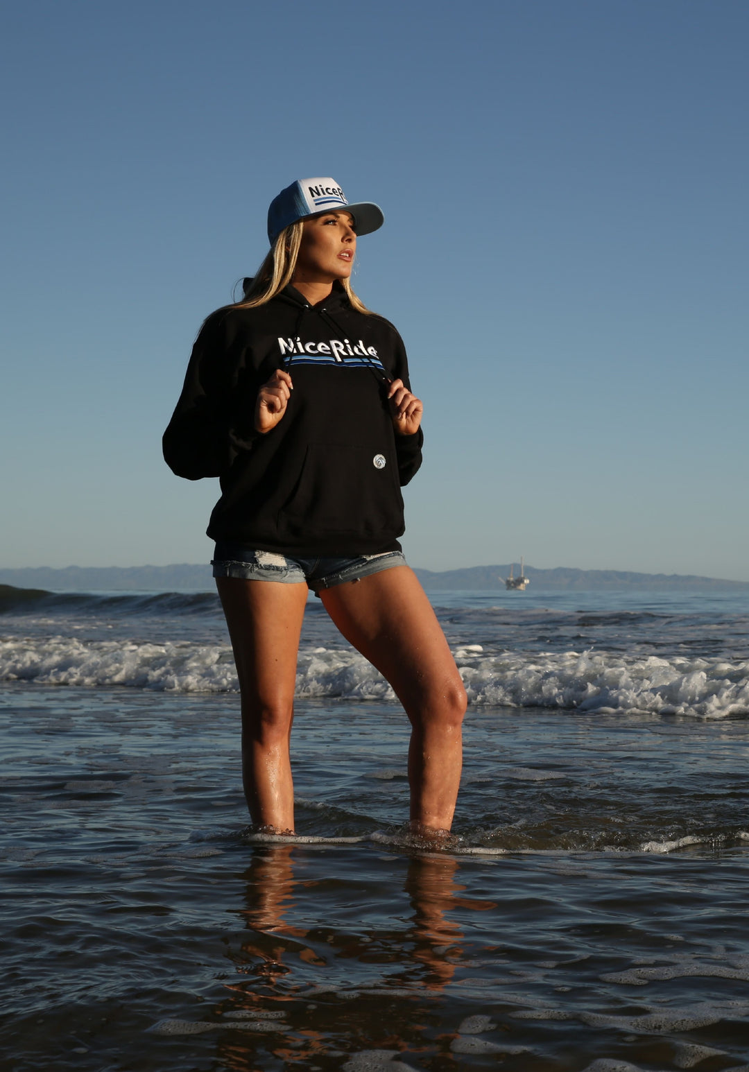 Woman wearing a black hoodie with NICERIDE logo standing in water, showcasing a stylish hoodie by the beach.